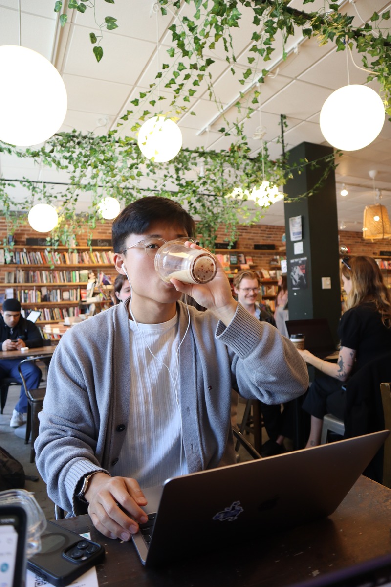 A man with glasses at a café table with a laptop, beneath hanging lights and greenery.