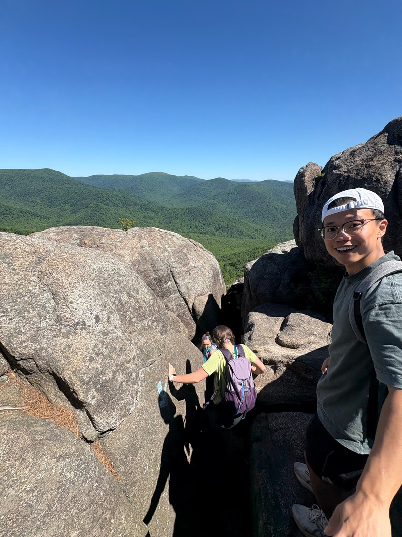 A hiker standing beside a large rock, with green mountain ridges behind him.