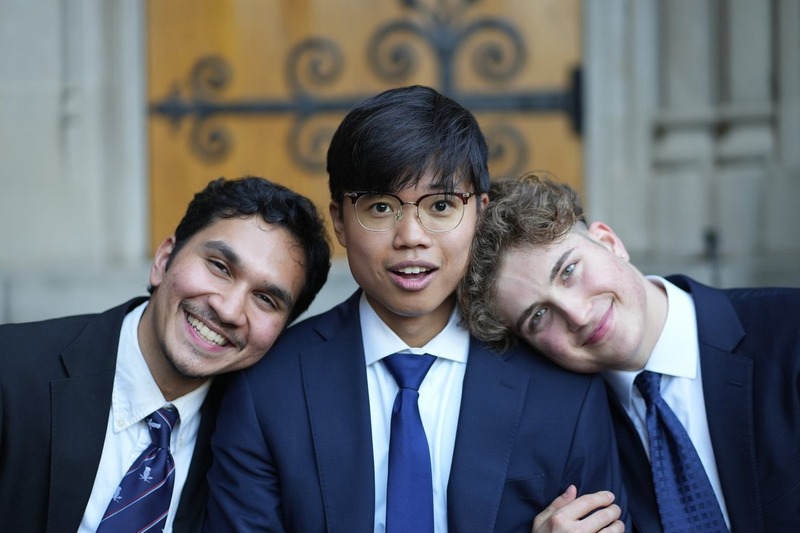 Three people in suits leaning together in front of an arched doorway.