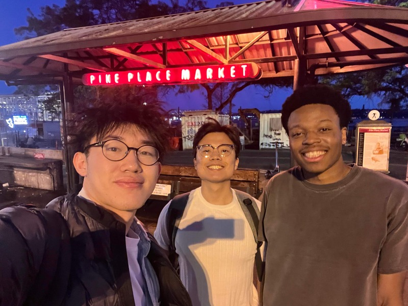 Three people beneath the illuminated Pike Place Market sign at night.