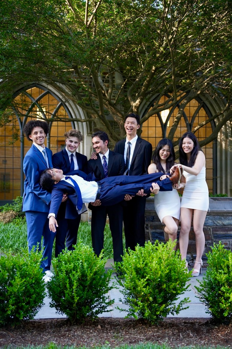 Friends in formal clothes holding a man horizontally in front of arched windows and a leafy tree.