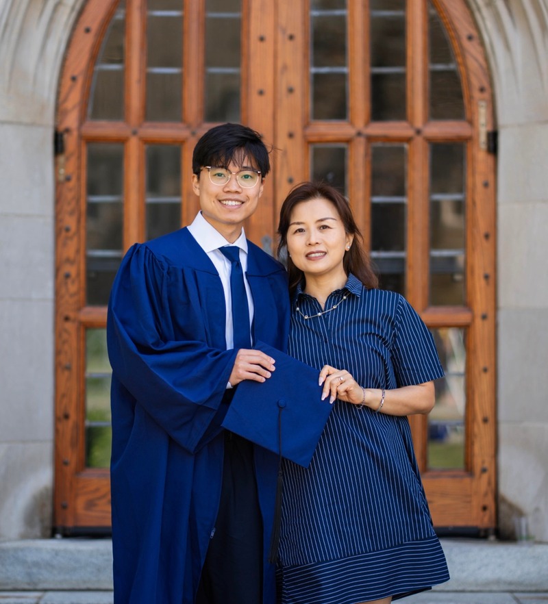 A graduate in a blue gown standing with a woman in front of a wooden chapel door.