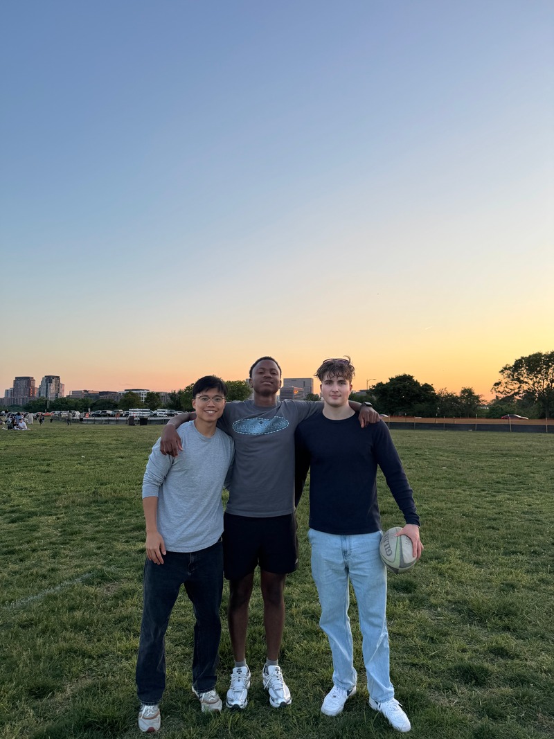 Three friends standing arm in arm on a grassy field at sunset, one holding a basketball.
