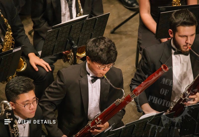 A musician wearing glasses and formal concert clothes, holding a bassoon among an orchestra.